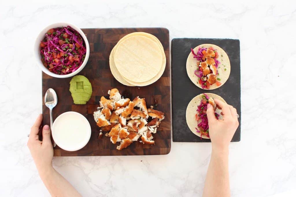 Two hands assembling fish tacos on brown cutting board.