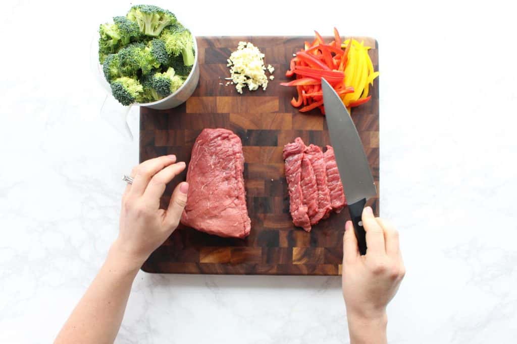 Two hands cutting beef on brown cutting board.