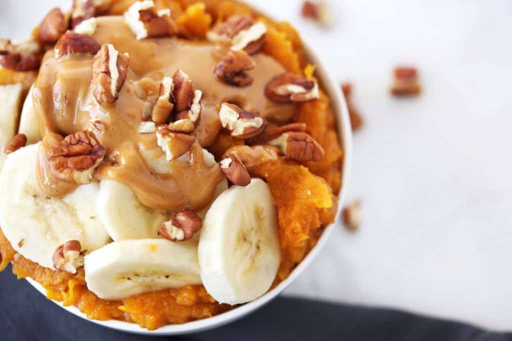 Sweet potatoes with banana slices, peanut butter and walnuts in a white bowl on grey napkin.
