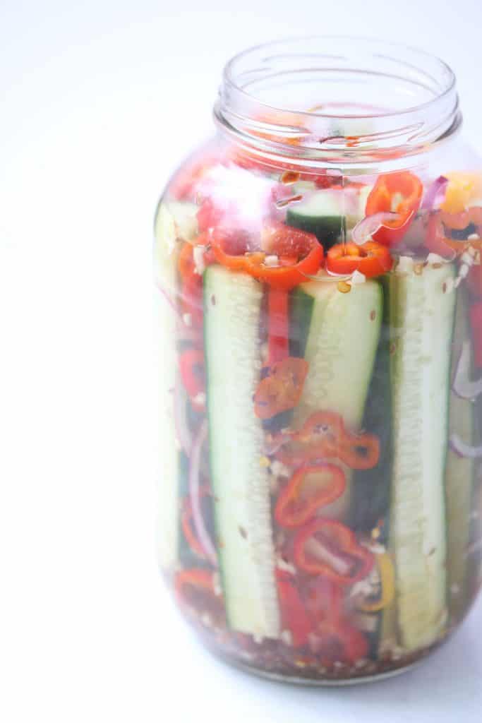 Pickles and peppers in clear mason jar on white surface.