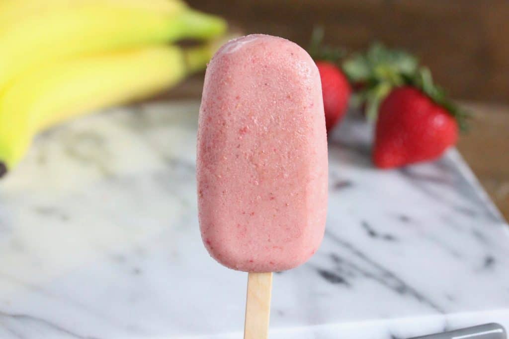 Pink popsicles on marble surface next to strawberries and bananas.