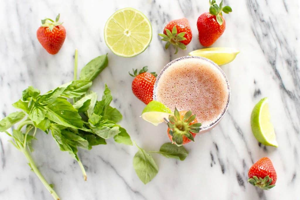 Pink margarita next to strawberries, basil and limes.