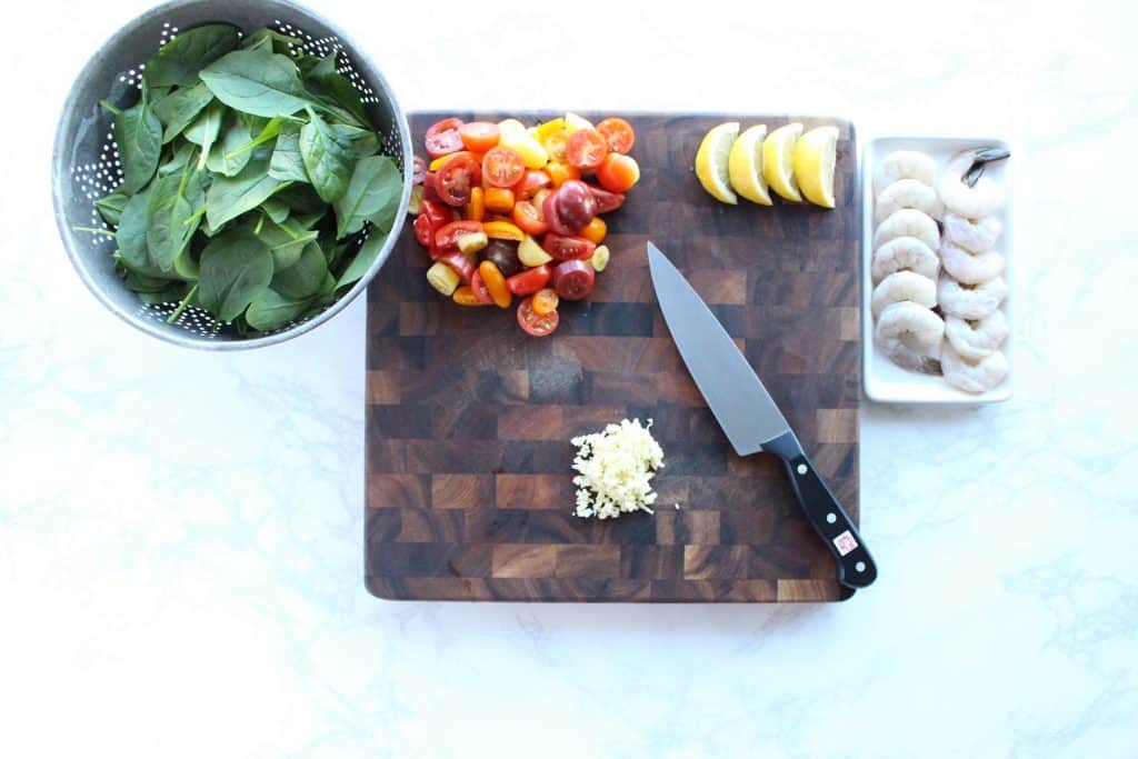 Spinach, tomatoes, lemons, garlic and shrimp on brown cutting board next to chef's knife.