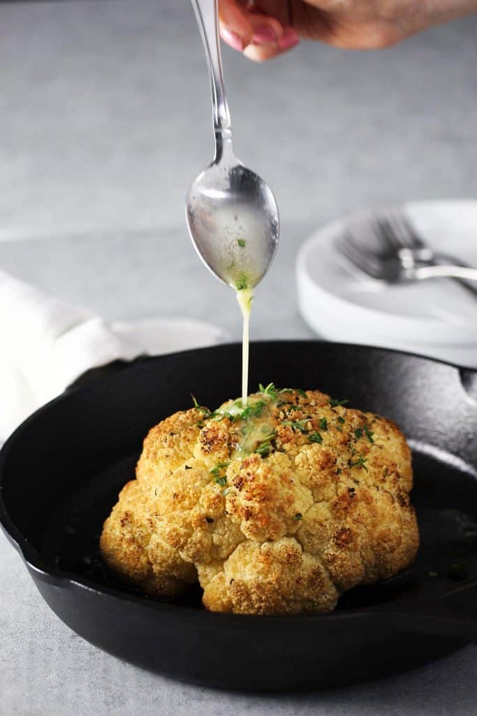 Whole head of roasted cauliflower in cast iron pan with butter being poured on top.